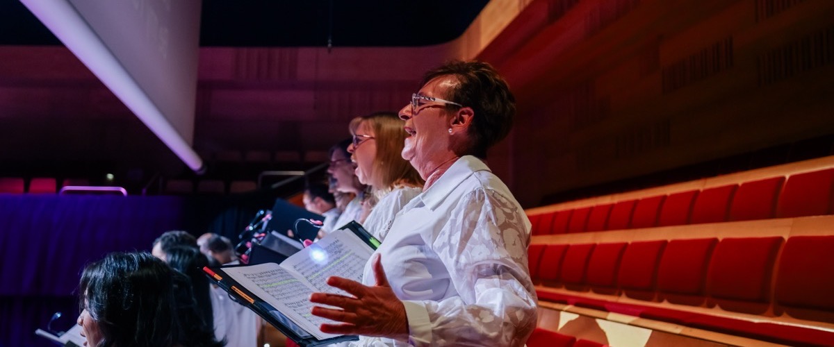 A group of people singing in an auditorium, with one person prominently holding sheet music.