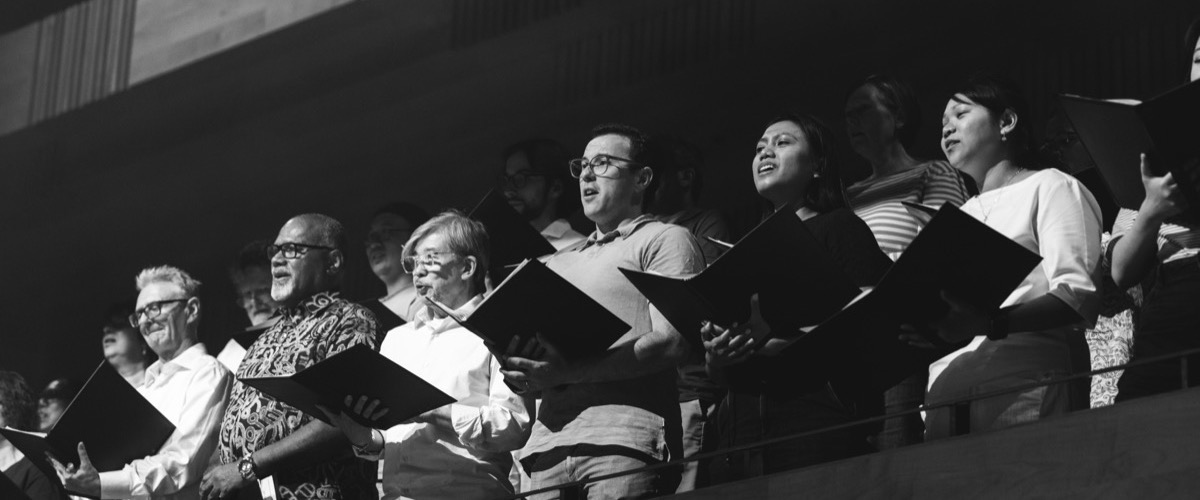 A group of people, some wearing glasses, sing while holding sheet music and standing in a choir.