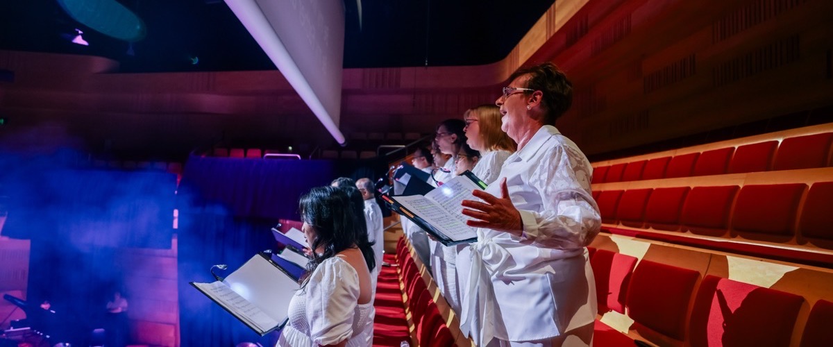 A group of people in a choir, dressed in white, sing in a theater with red seats.