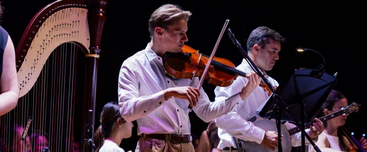 A group of musicians play instruments on stage, with a harp, violin, and banjo visible.