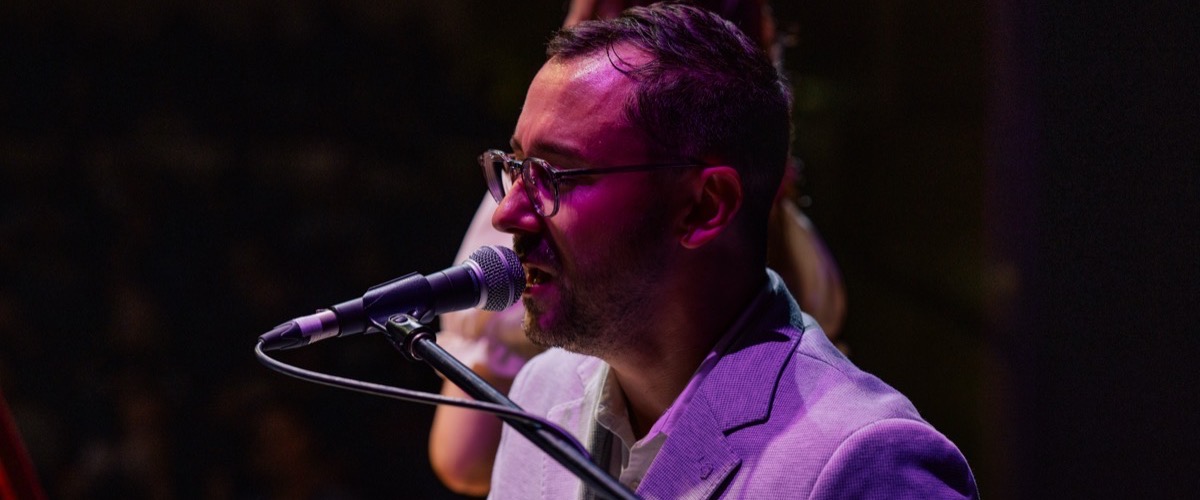 A man with short dark hair and glasses sings into a microphone on a stand.