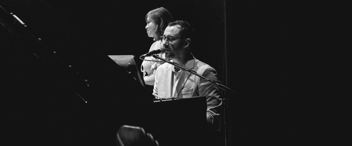 This black-and-white photograph depicts two individuals seated at a microphone stand, each holding a sheet of paper.