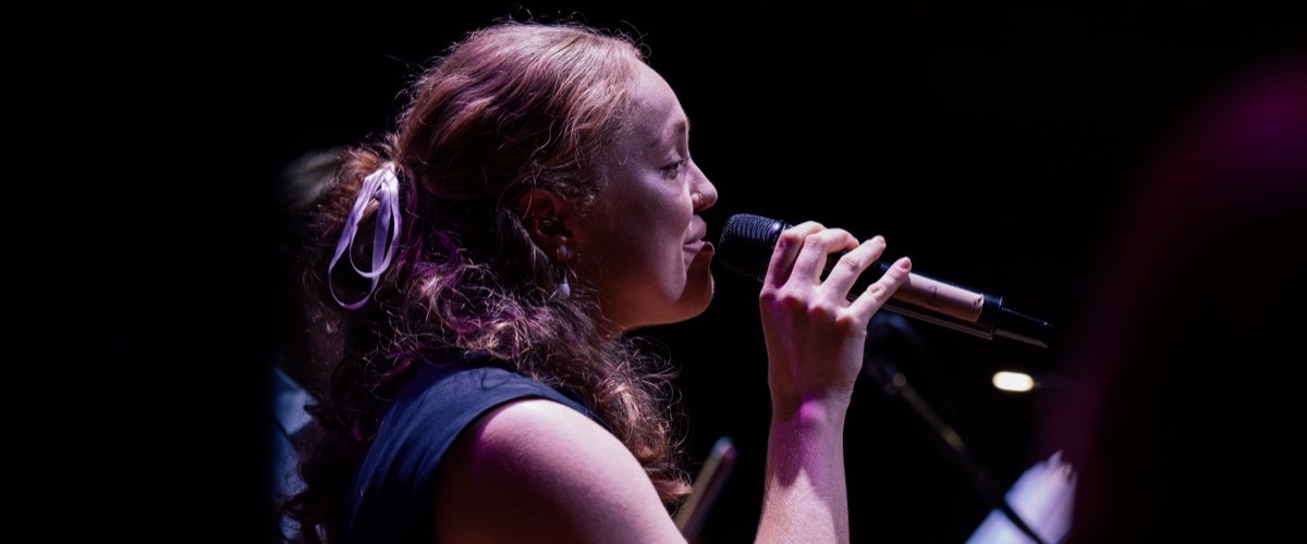 A woman with long, curly hair and a bow in her hair sings into a microphone.
