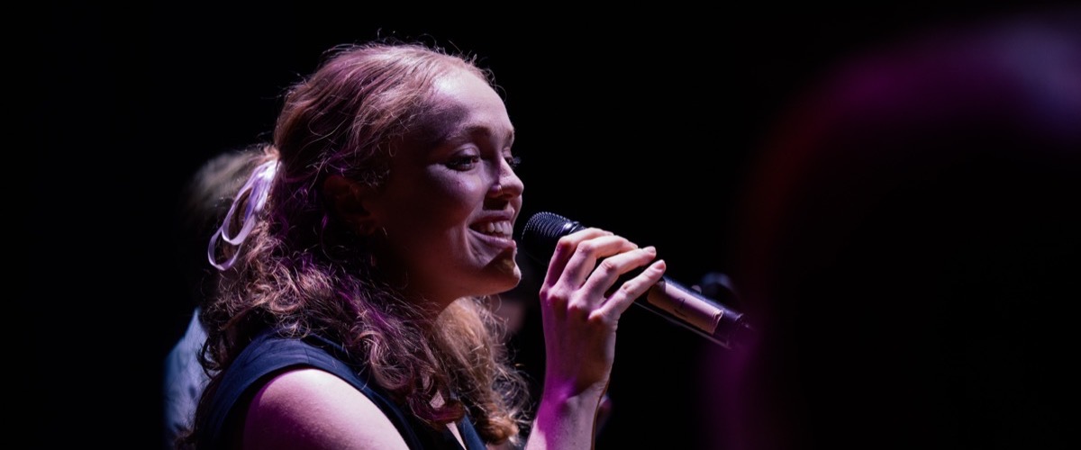 A woman singing into a microphone, set against a dark background with a purple light shining on her.