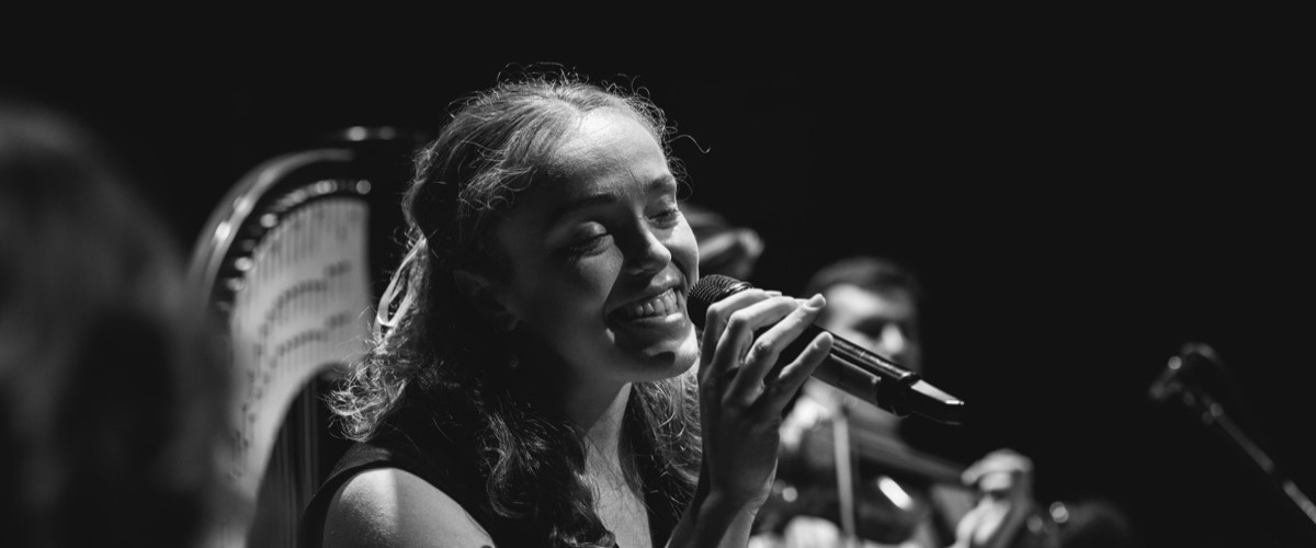 A woman with long hair sings into a microphone, accompanied by a harpist in the background.