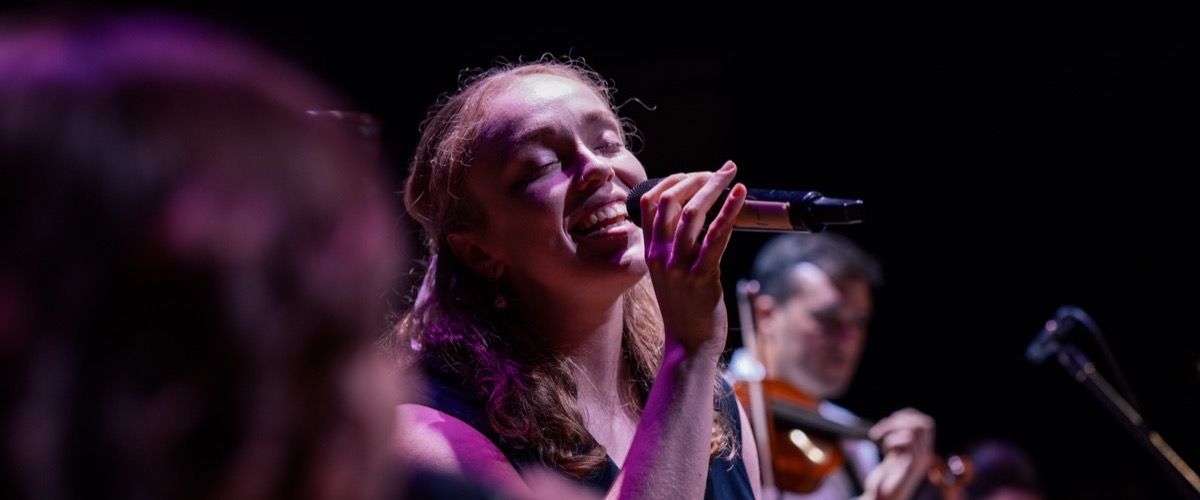 A woman sings into a microphone on stage, accompanied by a musician playing a violin.