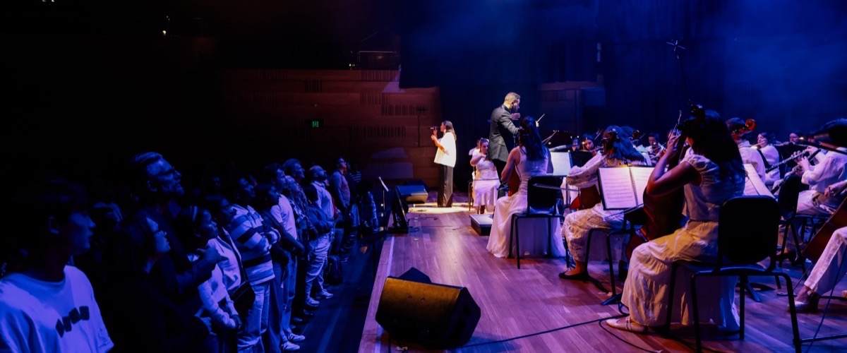 A group of musicians and audience members are performing on a stage, illuminated by blue light.