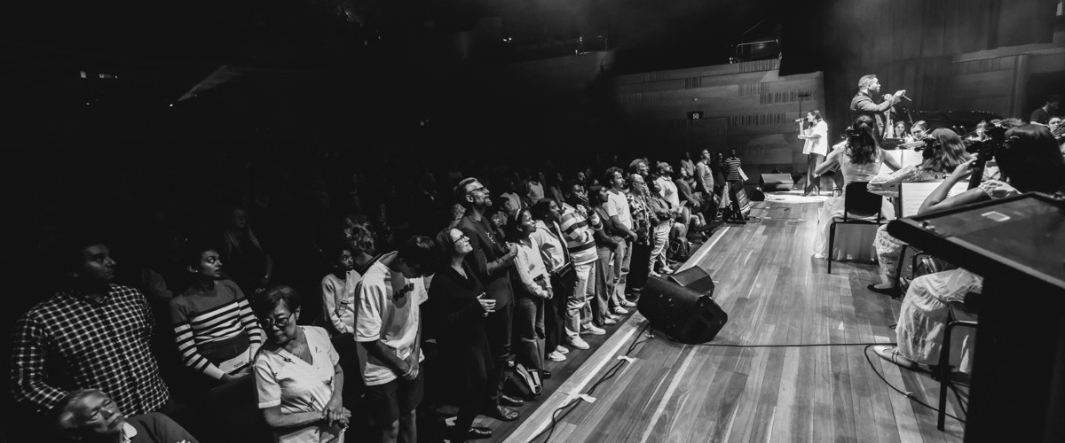 Graph depicts a group of people standing on a stage, facing a seated audience, with some individuals playing musical instruments.