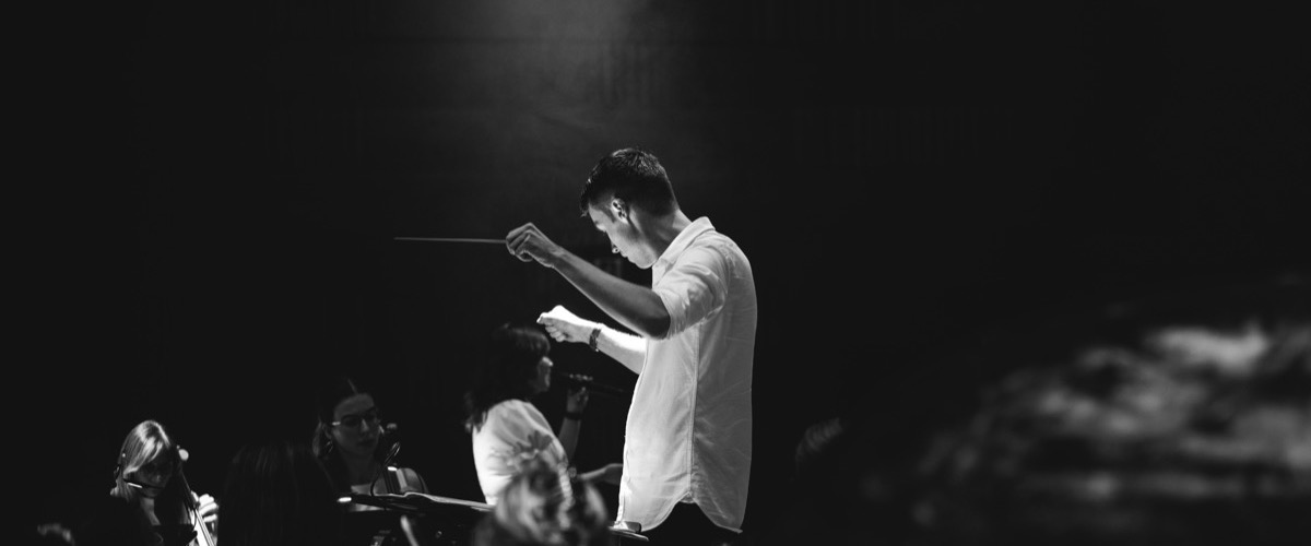 A conductor leads an orchestra, holding a baton and sheet music, in a dimly lit room with a spotlight shining down.