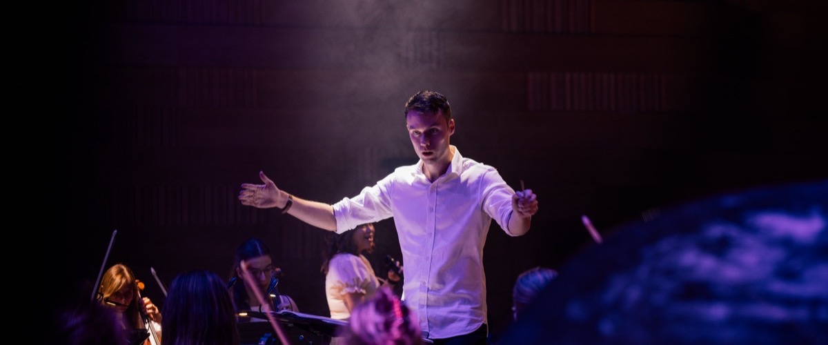 A man stands in front of an orchestra, his arms outstretched and hands open.