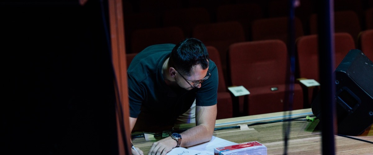A man is leaning over a table in an auditorium, surrounded by papers and other objects.