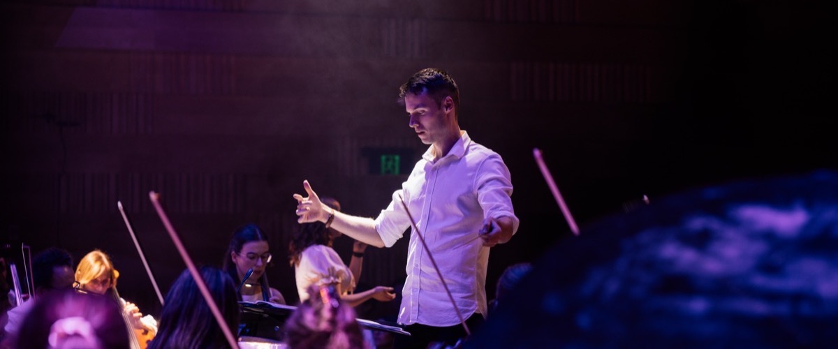 A conductor leads an orchestra, standing with arms outstretched and pointing at musicians.