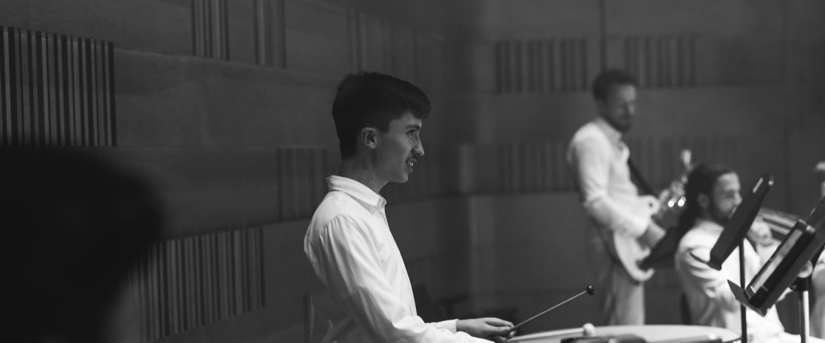A group of musicians performing in a recording studio, with one person playing a drum set in the foreground.