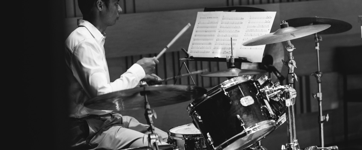 A man in a white shirt plays a drum set, with sheet music on a stand nearby.