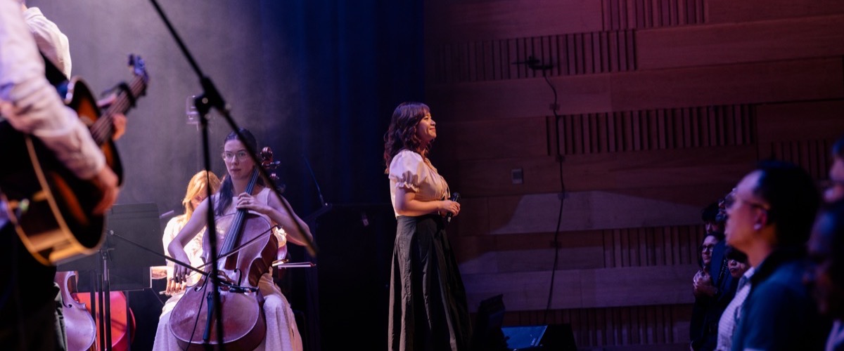 The photograph depicts a group of musicians performing on stage, with a cellist in the foreground and a singer in the background.