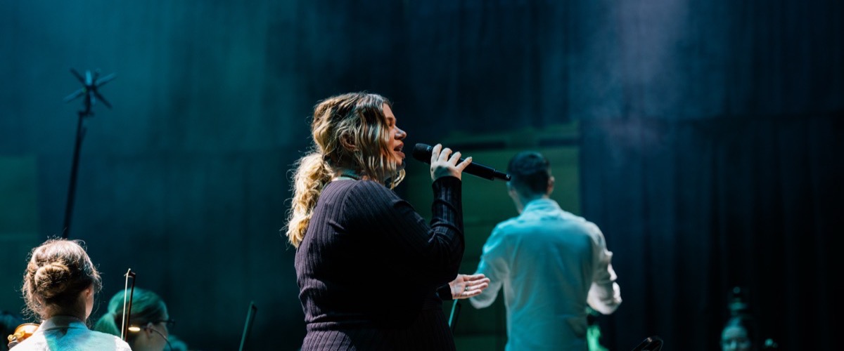 A woman singing into a microphone on a stage with an orchestra playing in the background.