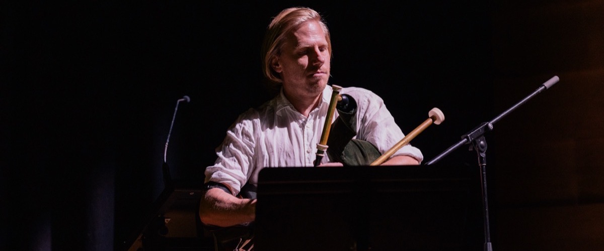 A man seated on a chair, holding a guitar and a stick-like instrument.