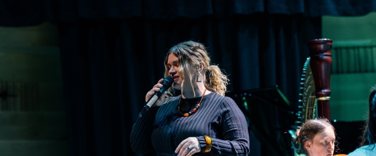 A woman is singing into a microphone on stage, accompanied by a harp and another person.