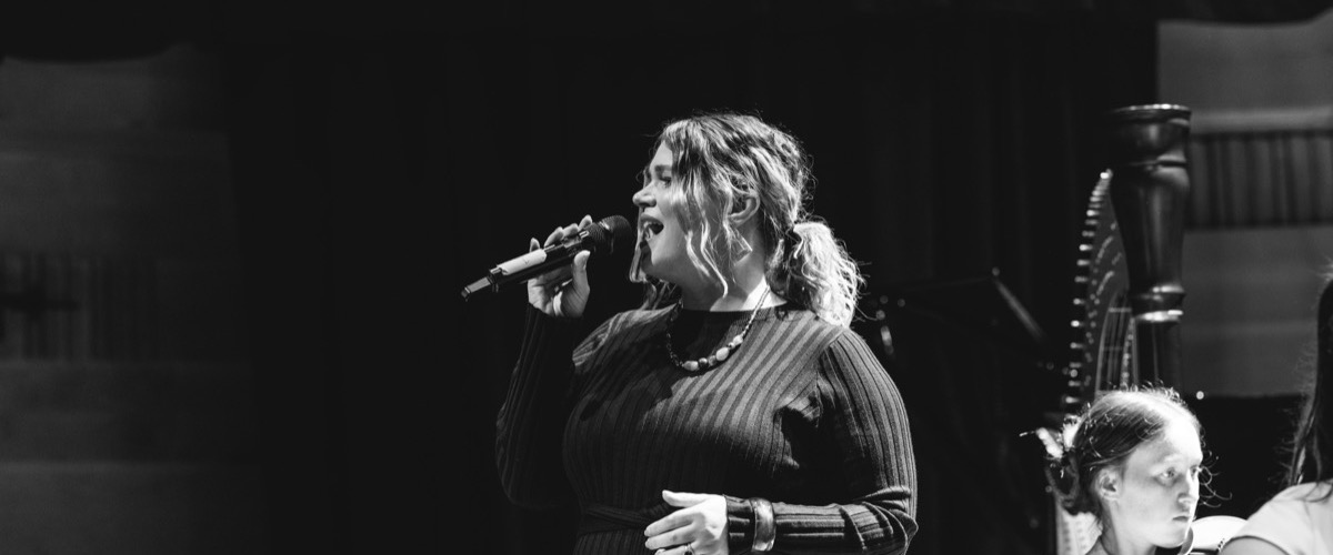 A woman sings into a microphone, accompanied by two individuals, in a setting that appears to be a stage or performance area.