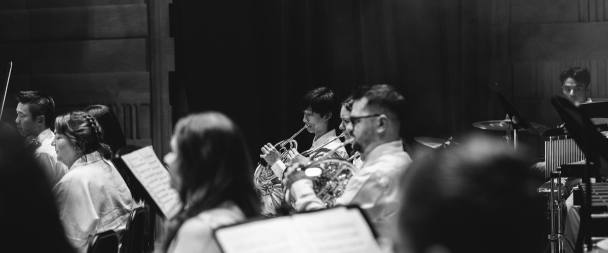 A group of musicians in a band are performing in a dimly lit room, likely a concert hall.