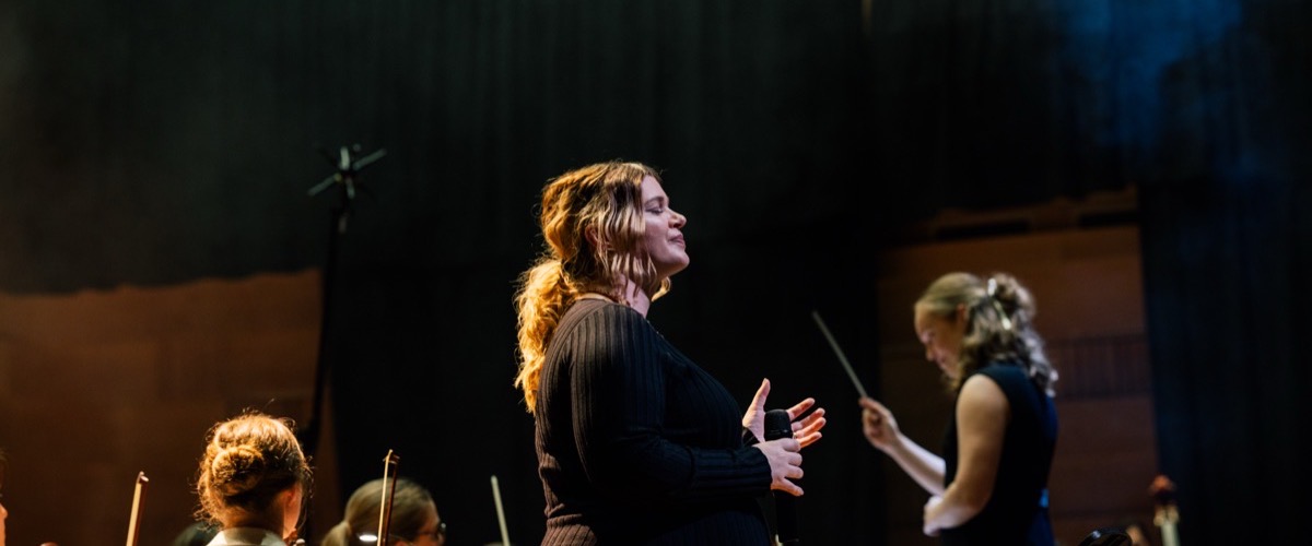 The photograph depicts a woman singing in front of a group of musicians in an orchestra setting.
