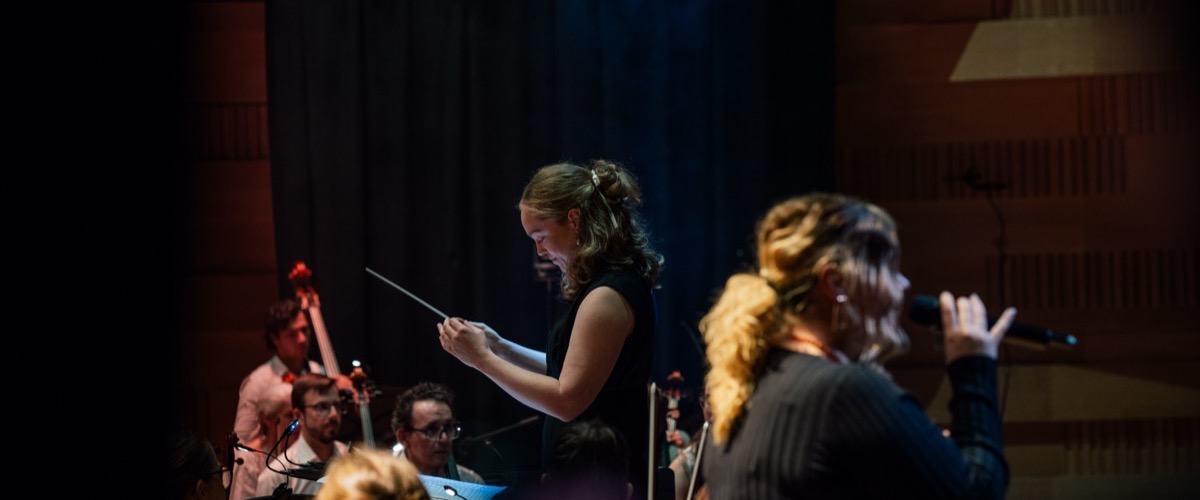 Graph depicts a woman holding a conductor's baton and standing in front of a group of musicians.