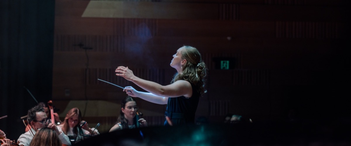 A woman, likely a conductor, stands in front of an orchestra, holding a baton in her right hand.
