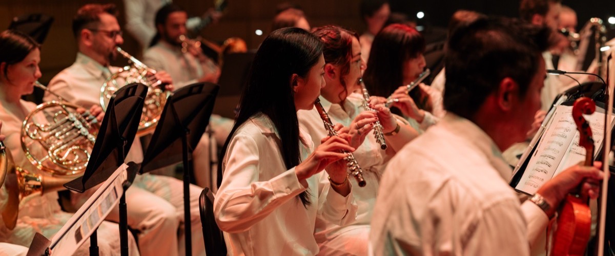 A group of musicians are playing instruments, likely in an orchestra, with a focus on brass and woodwind instruments.