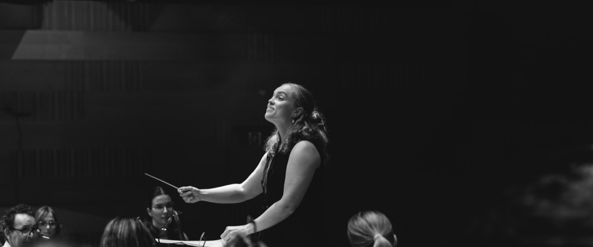 The photograph depicts a woman conducting an orchestra, holding a baton in her right hand.