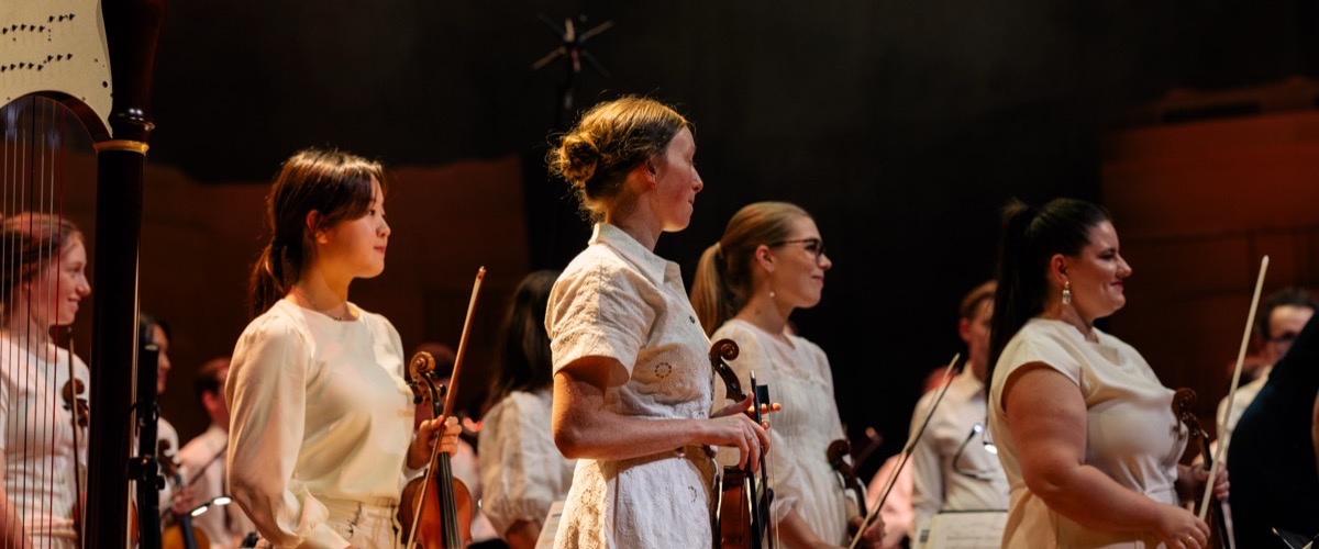 The photograph shows a group of women standing in a line, each holding a violin or harp.