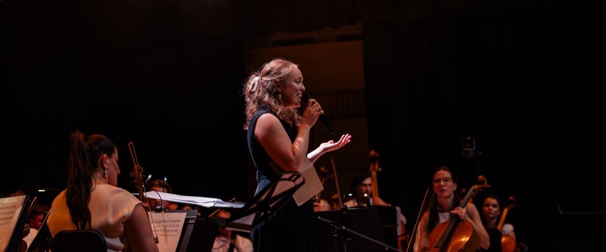 A woman singing into a microphone on stage with a group of musicians playing string instruments.