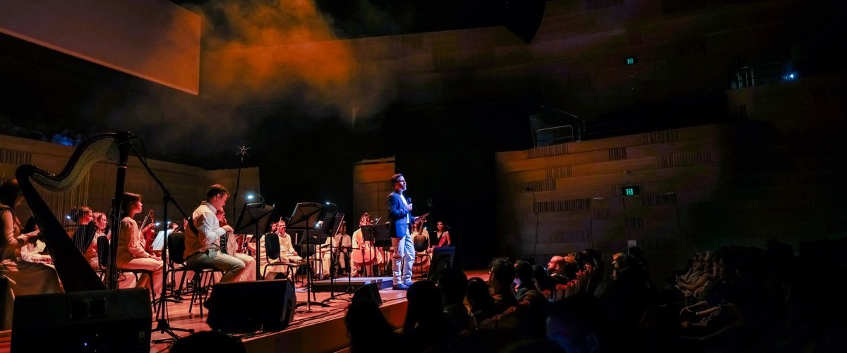 A man in a blue suit addresses an audience on stage, accompanied by an orchestra seated behind him.