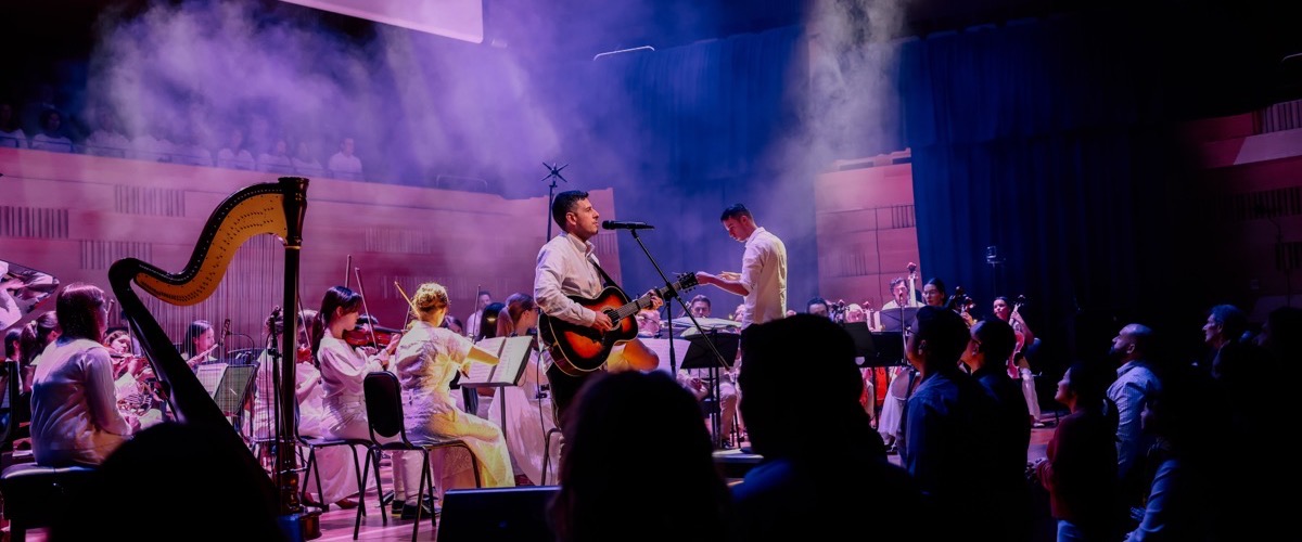 A group of musicians, including a harpist and guitarist, perform on stage in front of an audience.