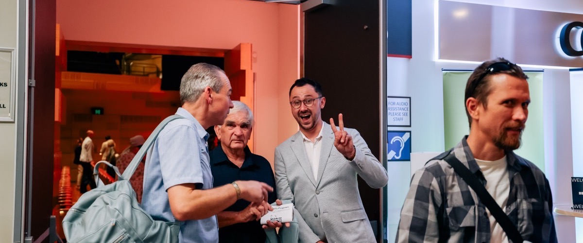 Four men stand in a doorway, with one man holding a small white box and another making a peace sign with his hand.