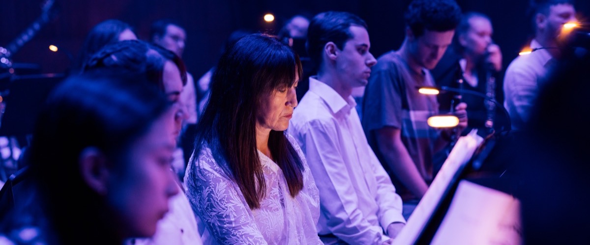 A group of people are gathered in a dimly lit room, seated and facing the same direction.