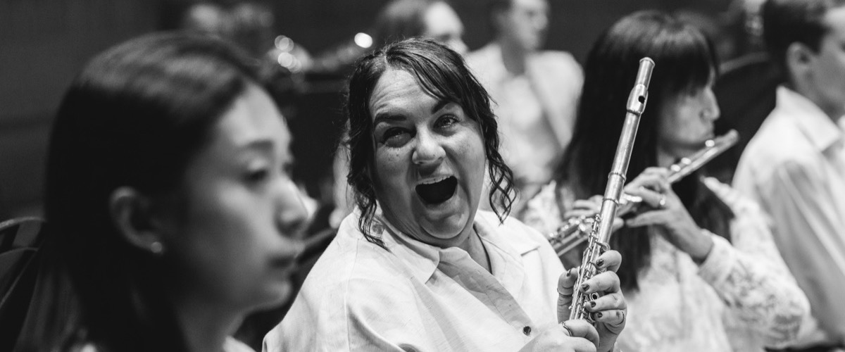 This black-and-white photograph depicts a woman holding a flute, surrounded by a group of people playing instruments.
