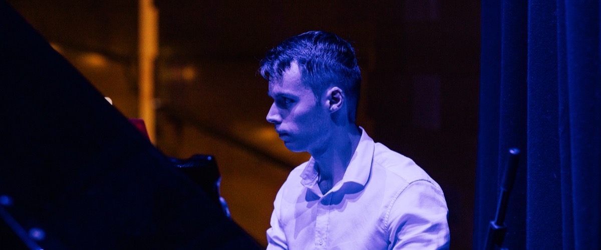 A man in a white collared shirt standing beside a grand piano.