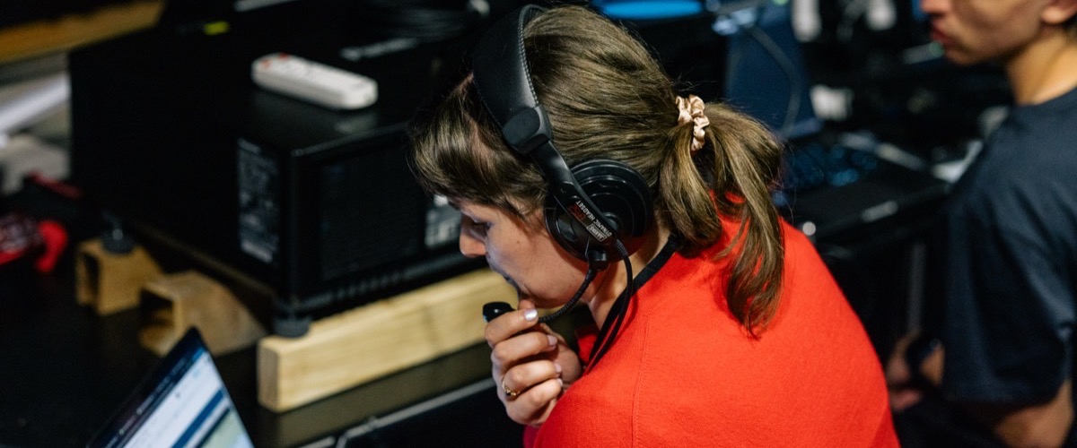 The photograph depicts a woman wearing a headset and holding a microphone, with a man standing in the background.