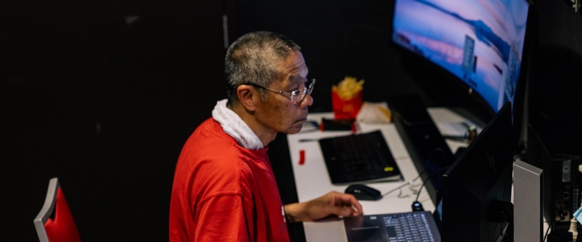 A man in a red shirt sitting at a desk with a computer monitor and keyboard.