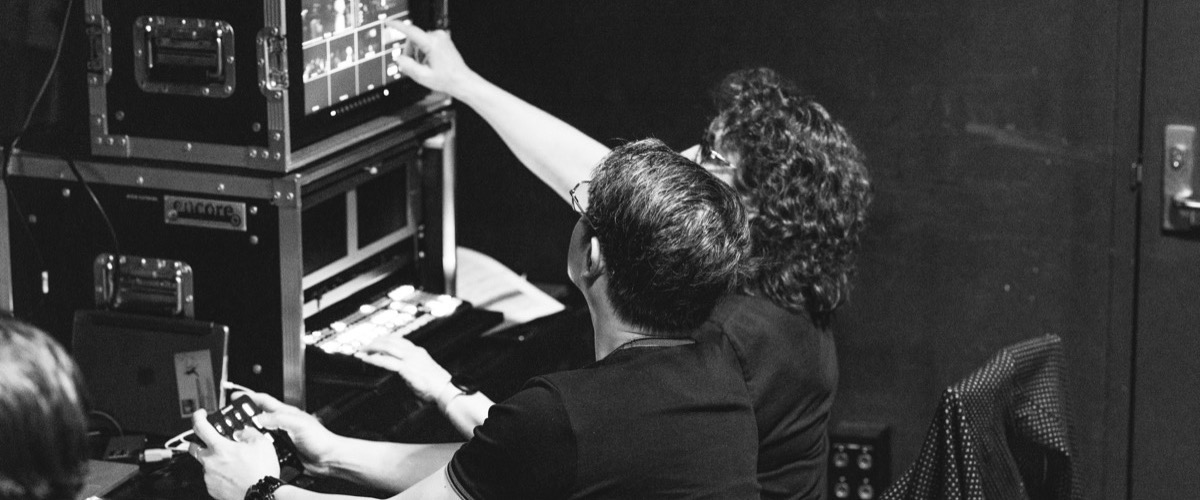 This black-and-white photograph depicts two people working in a control room, with one person operating a video switcher and the other monitoring a screen.