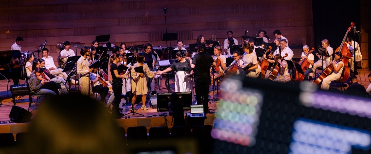 A group of musicians performing on stage, with instruments and music stands in view.