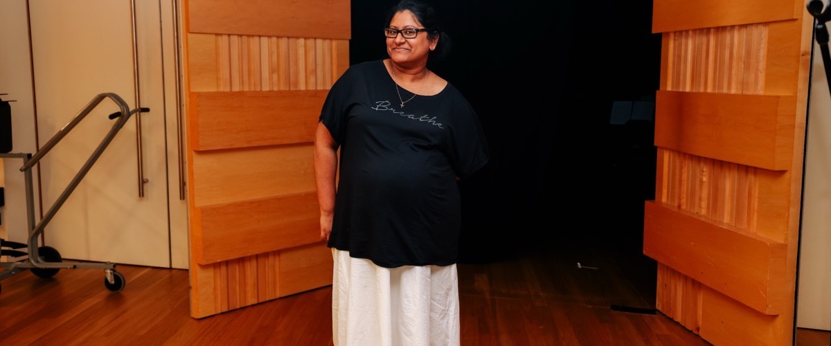 A woman stands in a room with wooden doors and a metal frame on wheels, wearing a black shirt and white pants.