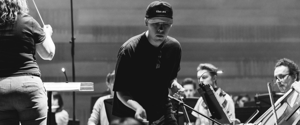 This black and white photograph depicts a man in a dark shirt and hat, holding a guitar, standing in front of an orchestra.