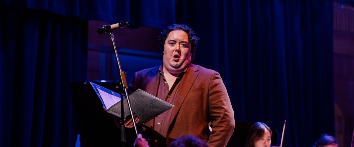 A man in a suit holds a music stand with sheet music, accompanied by a microphone and an orchestra in the background.