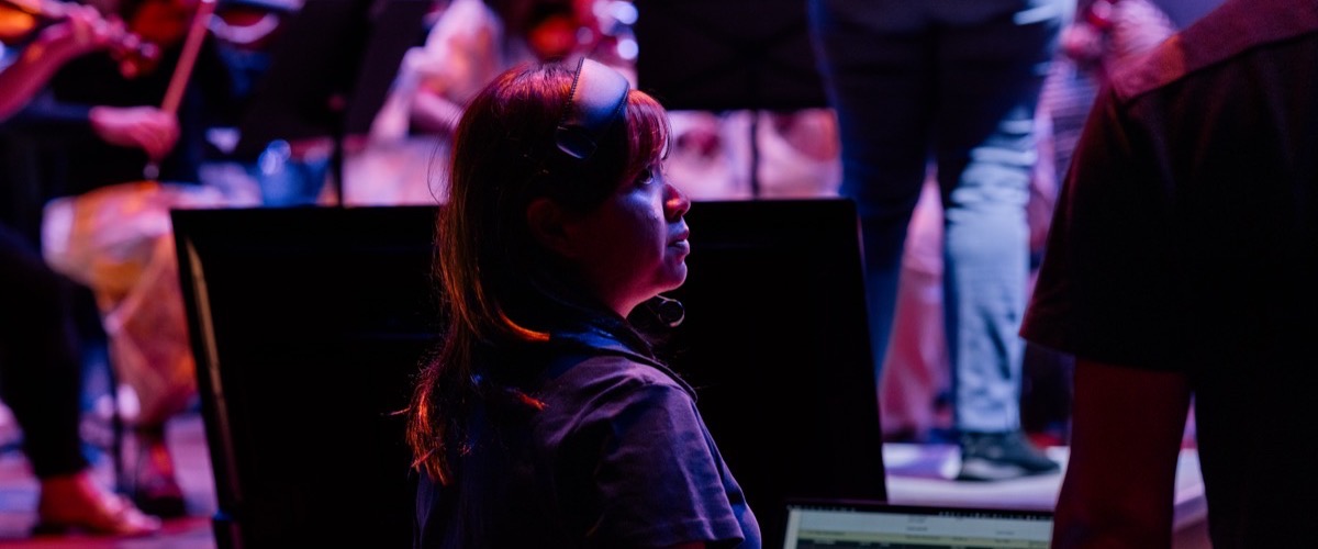 The photograph depicts a woman wearing headphones in a dimly lit room with people playing instruments in the background.