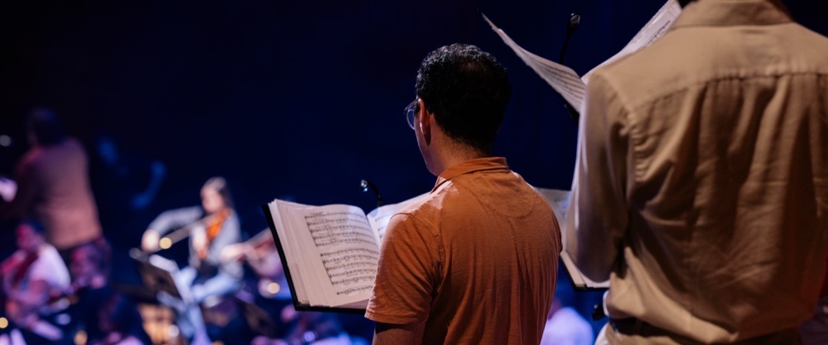 A group of people, likely a choir, are rehearsing on a stage, with some holding sheet music and others playing instruments.