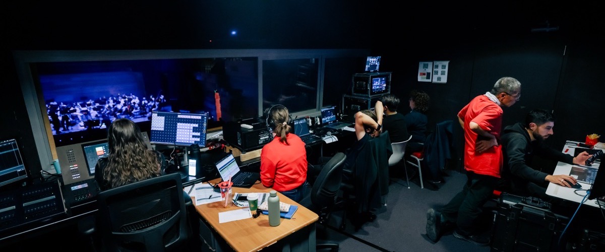 A darkened control room with a group of people seated at desks, facing a large video wall.