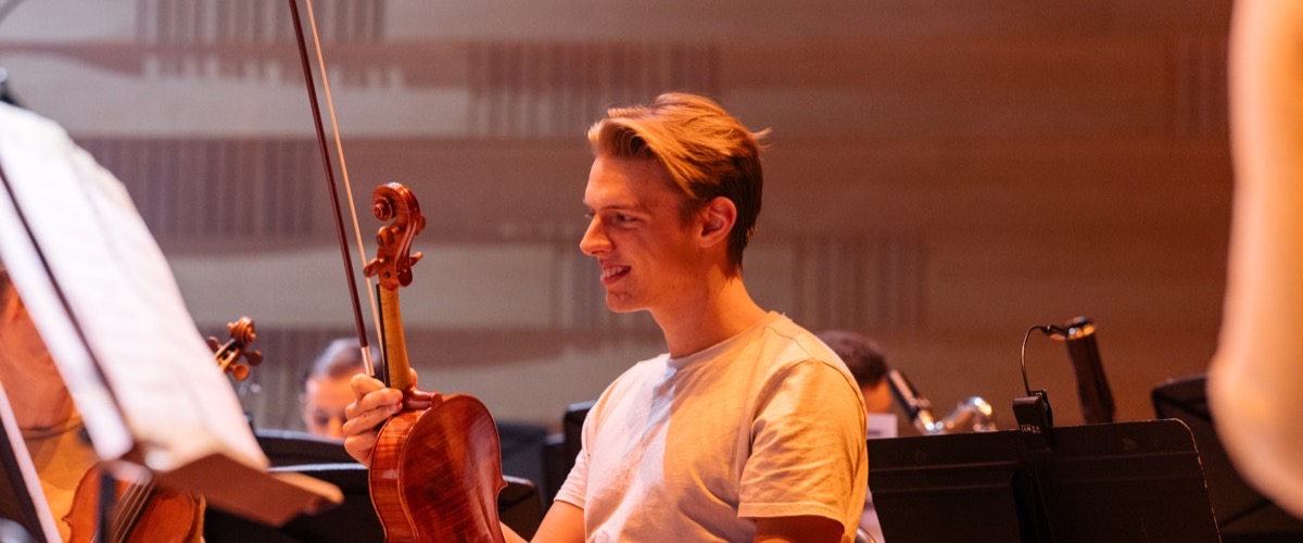 Graph depicts a man holding a violin in a concert hall setting, surrounded by other musicians.