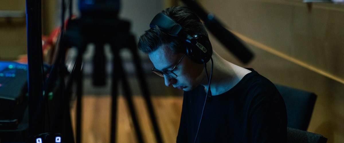 A man wearing headphones in a dimly lit room with a tripod and other equipment in the background.