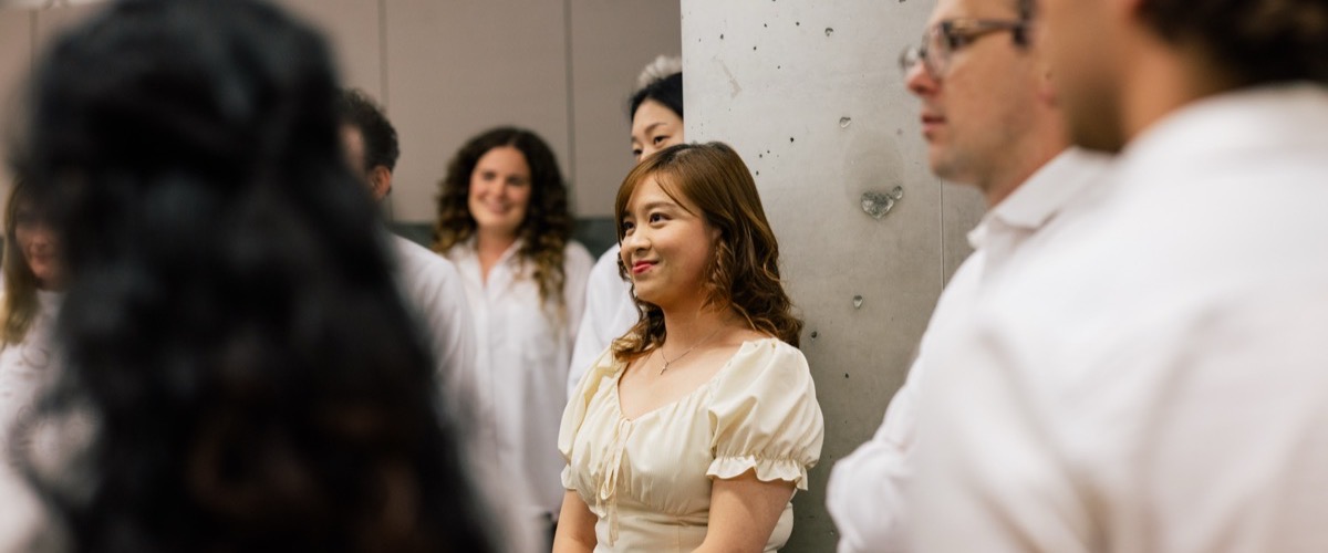 A woman in a cream-colored top stands in front of a group of people, all wearing white shirts.